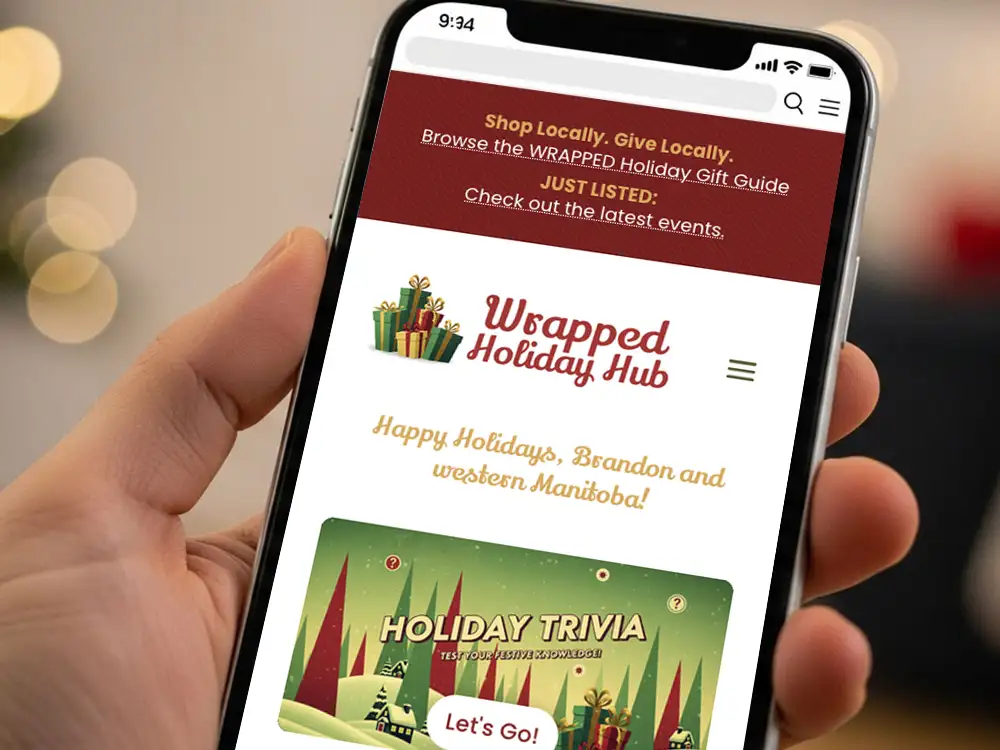 Close-up of a smartphone showing the home page of the Wrapped Holiday Hub website. Text says "Shop Locally. Give Locally. Browse the Wrapped Holiday Gift Guide. Just Listed: Check ou the latest events." The logo shows a stack of brightly coloured, wrapped gifts and the text "Wrapped Holiday Hub". A messages states "Happy Holidays, Brandon and western Manitoba!". At the bottom of the screen is a graphic for the "Holiday Trivia" section that reads, "Test your festive knowledge" with a button that reads, "Let's Go!".